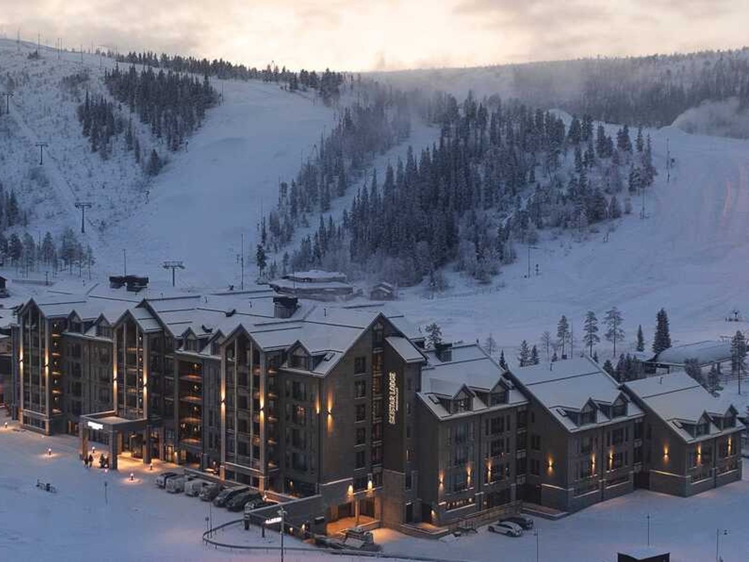 Et stort hotel med oplyste vinduer beliggende i en snedækket bjergskisportssted ved skumring.