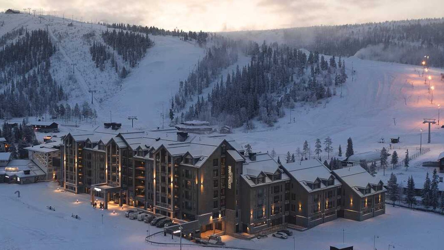 Et stort hotel med oplyste vinduer beliggende i en snedækket bjergskisportssted ved skumring.