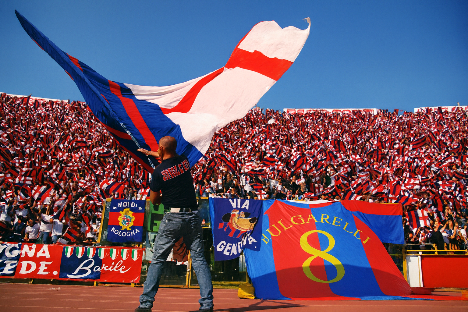 A man waves a large red, blue, and white flag on a stadium track, with stands packed with fans waving smaller flags behind him.