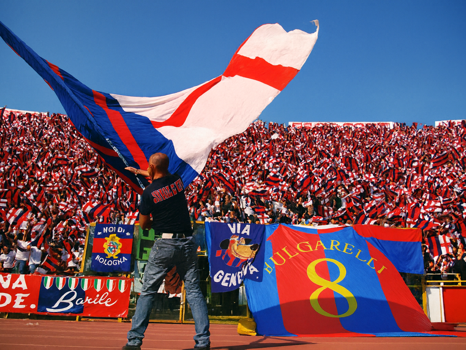 Un homme agite un grand drapeau rouge, bleu et blanc sur une piste de stade, avec des tribunes remplies de fans agitant de plus petits drapeaux derrière lui.
