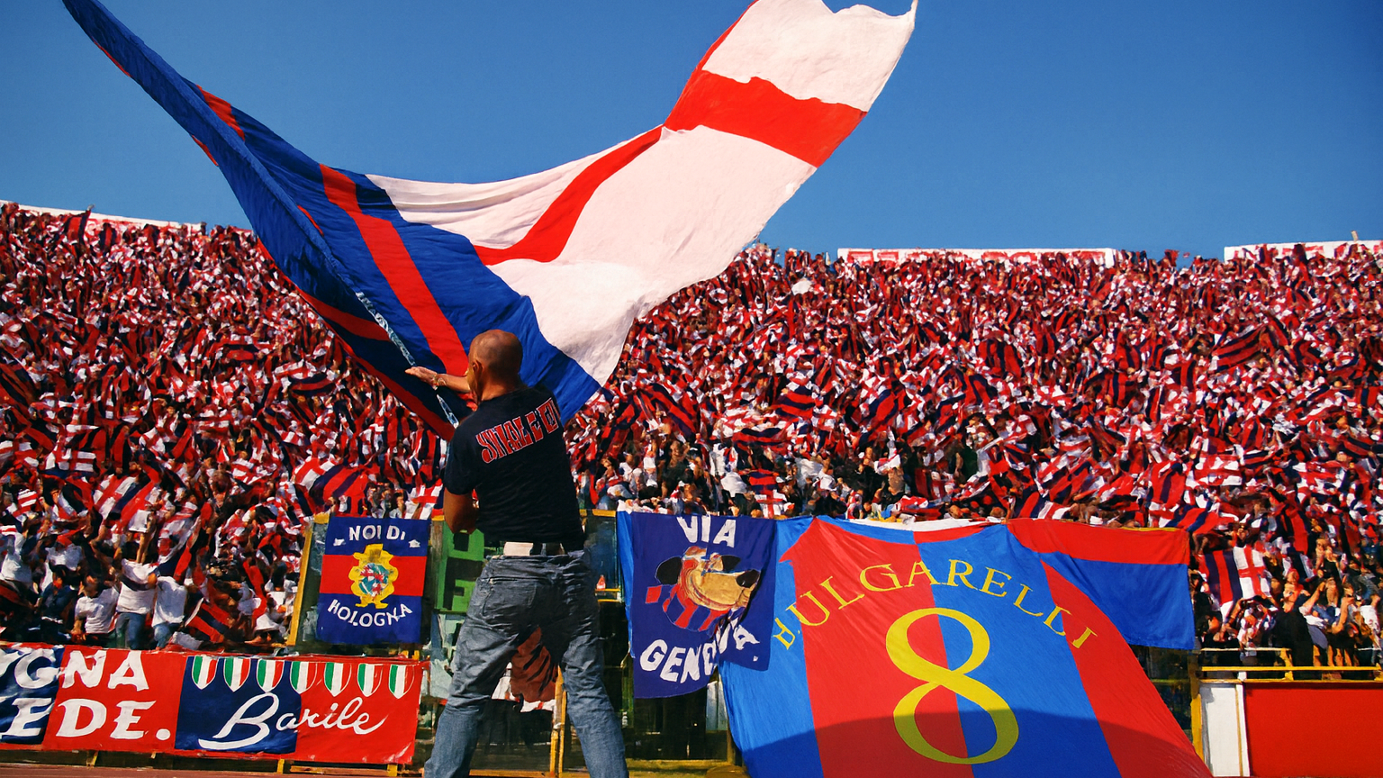 A man waves a large red, blue, and white flag on a stadium track, with stands packed with fans waving smaller flags behind him.