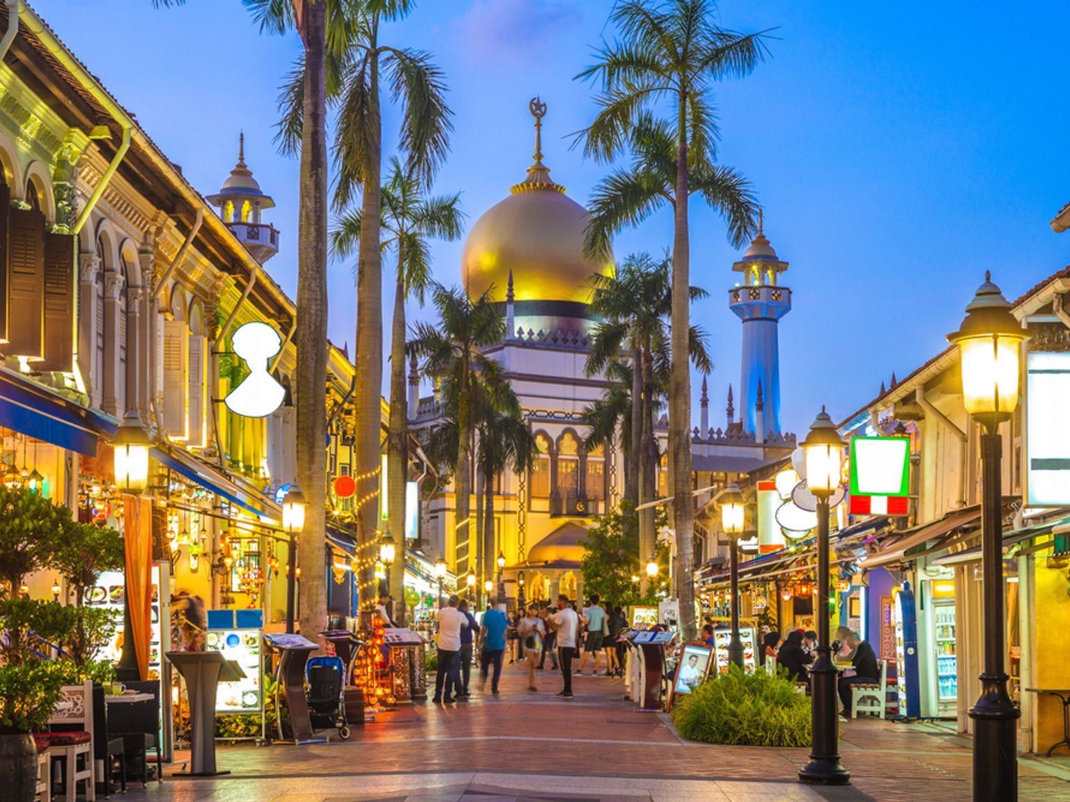 Oplyst gade med butikker og palmer, der fører til Singapores Sultan-moské med en gylden kuppel og minaret i skumringen.