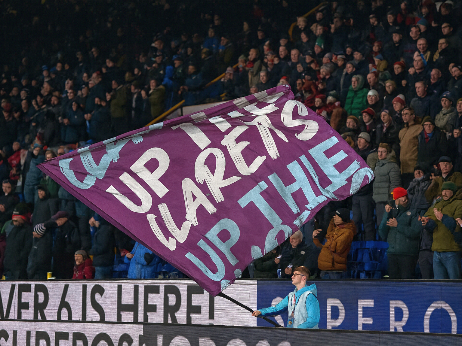 Un fan agite un drapeau violet avec l'inscription "UP THE CLARETS UP THE" en blanc, devant une foule de stade.