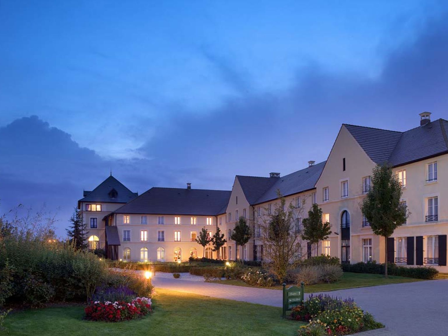 Flerbostadshus i krämfärg med upplysta fönster och mörka tak, belägna bland anlagda trädgårdar och stigar i skymningen. Hotel Campanile Val de France vid Disneyland® Paris