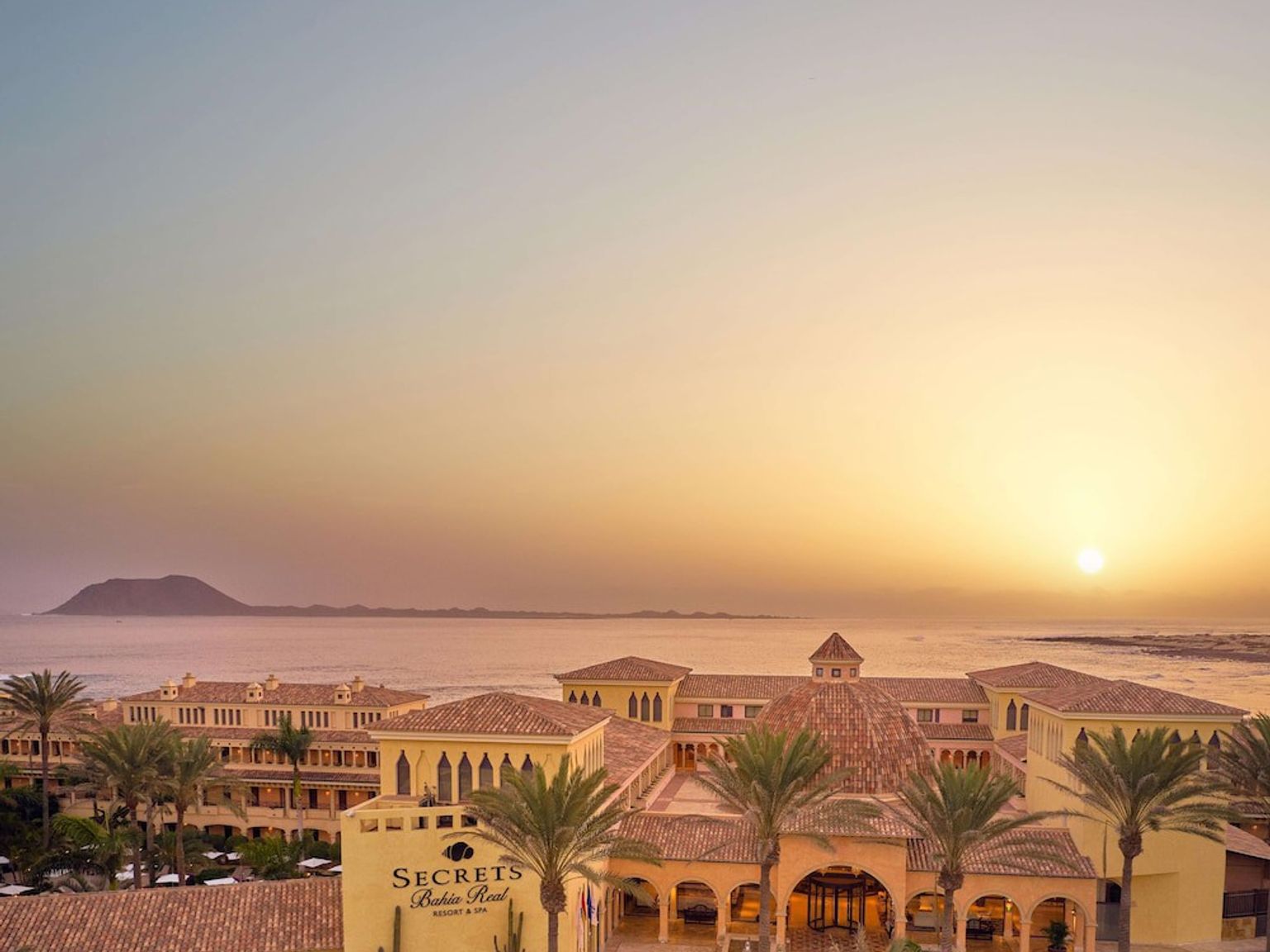 en luftvisning af en stor bygning med palmetræer foran havet ved solnedgang