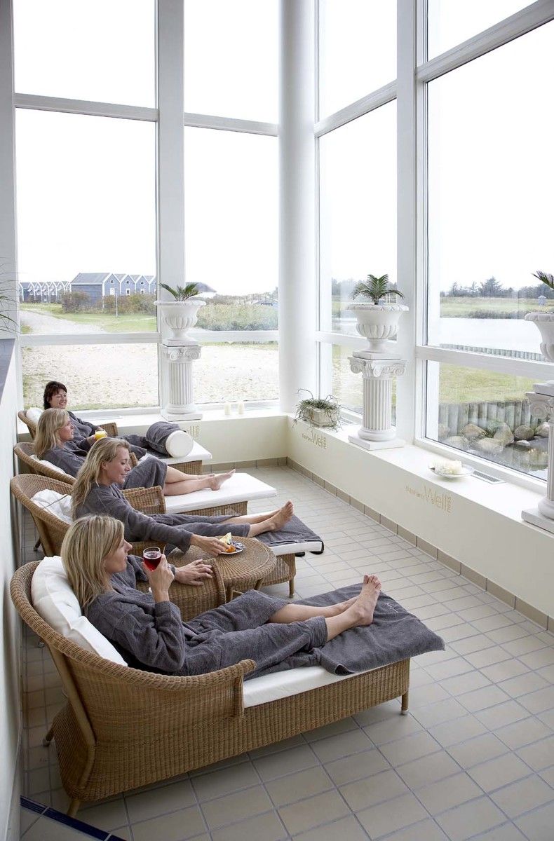 Flera kvinnor i grå badrockar kopplar av på vilstolar i ett ljust rum med stora fönster som vetter mot en utomhusvy.