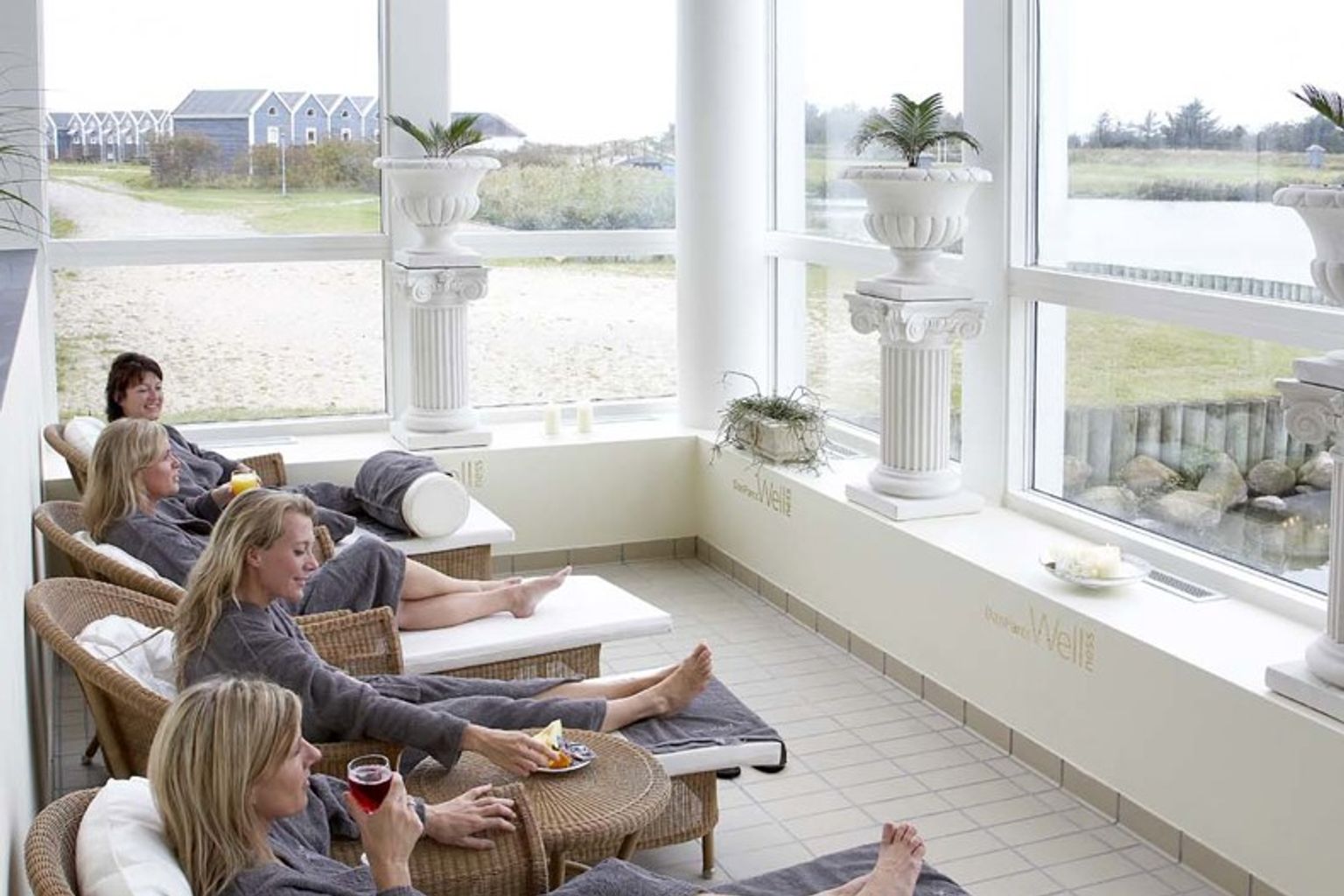 Flera kvinnor i grå badrockar kopplar av på vilstolar i ett ljust rum med stora fönster som vetter mot en utomhusvy.