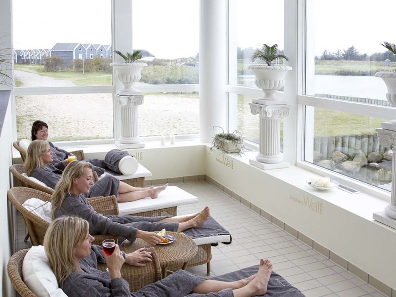 Flera kvinnor i grå badrockar kopplar av på vilstolar i ett ljust rum med stora fönster som vetter mot en utomhusvy.