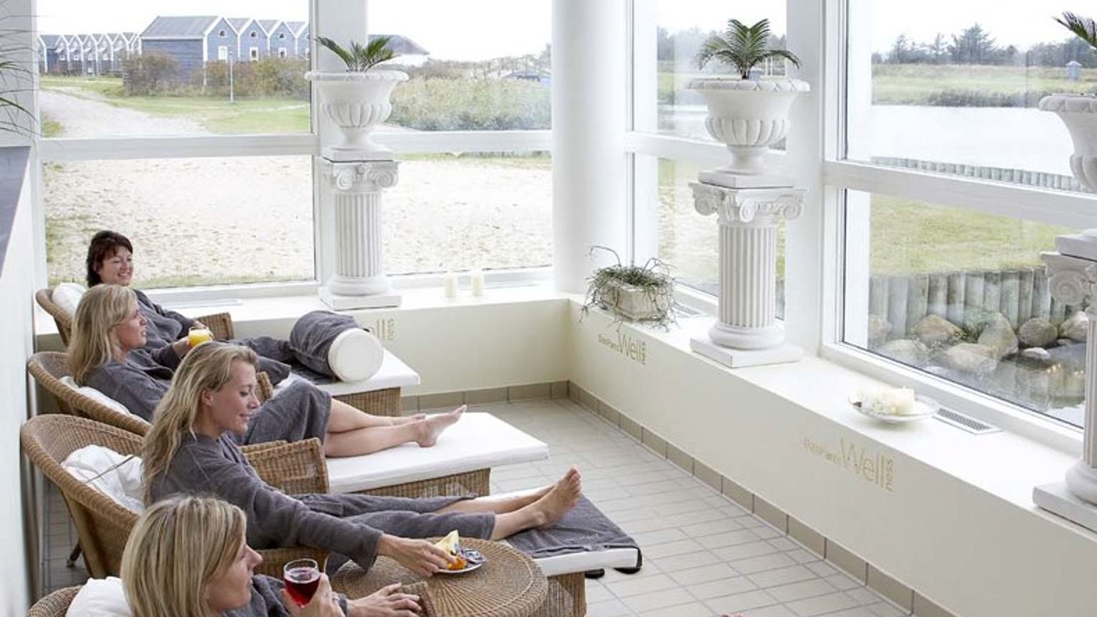 Flera kvinnor i grå badrockar kopplar av på vilstolar i ett ljust rum med stora fönster som vetter mot en utomhusvy.