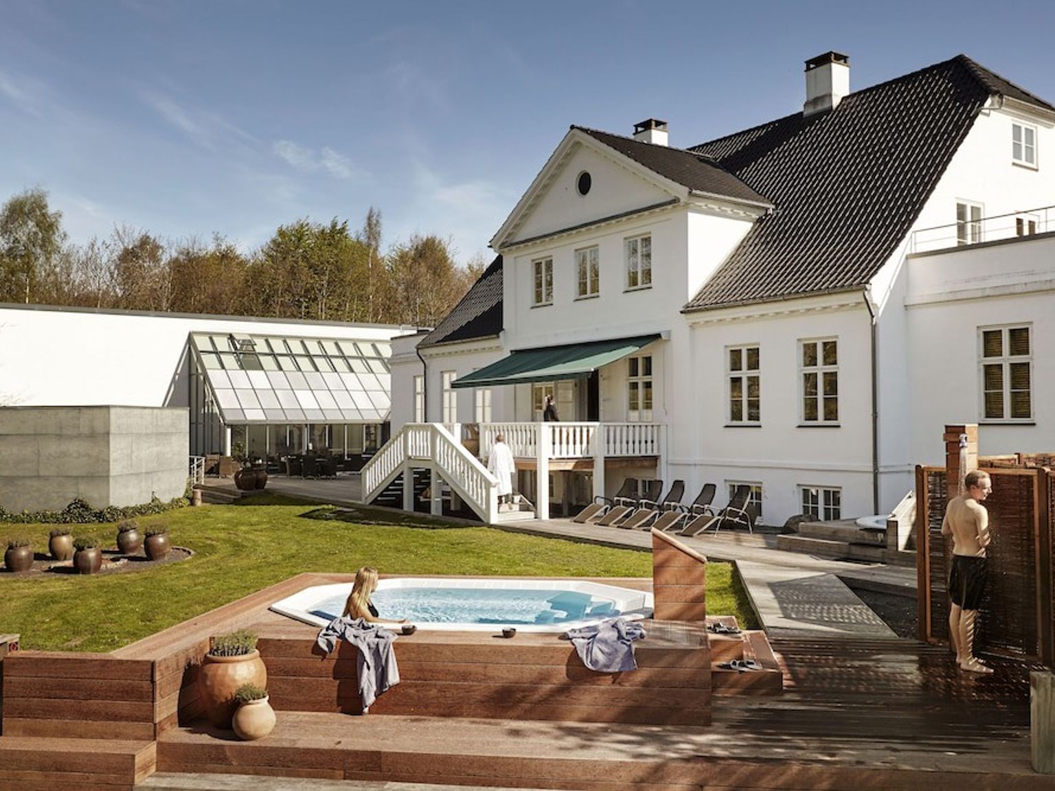 Ett utomhusspaområde med en stor vit byggnad, en bubbelpool på ett trädäck och en utomhusdusch, där människor njuter av faciliteterna under en klar himmel.