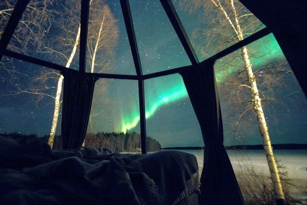 Grön norrsken strömmar över en stjärnklar himmel, sett från en säng i ett glastakrum med utsikt över en vinterskog och frusen sjö.