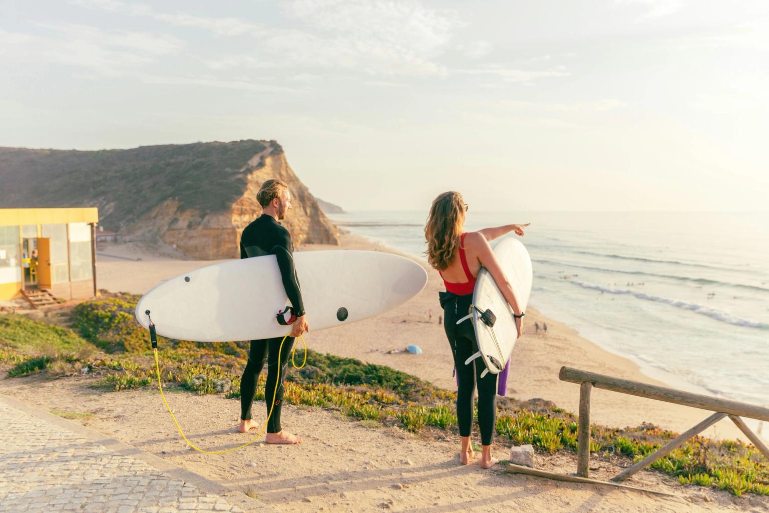 Två surfare i våtdräkter med brädor ser ut över en strand och hav vid solnedgången. Ericeira, Portugal