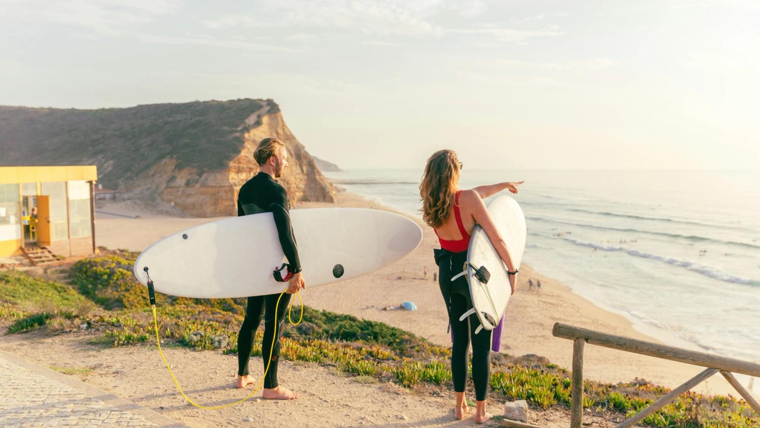 Två surfare i våtdräkter med brädor ser ut över en strand och hav vid solnedgången. Ericeira, Portugal