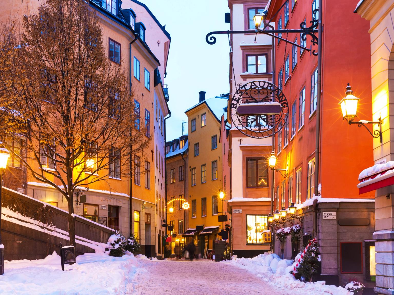 En snötäckt kullerstensgata i ett historiskt Stockholm, kantad med färgglada byggnader och lysande gatlyktor.