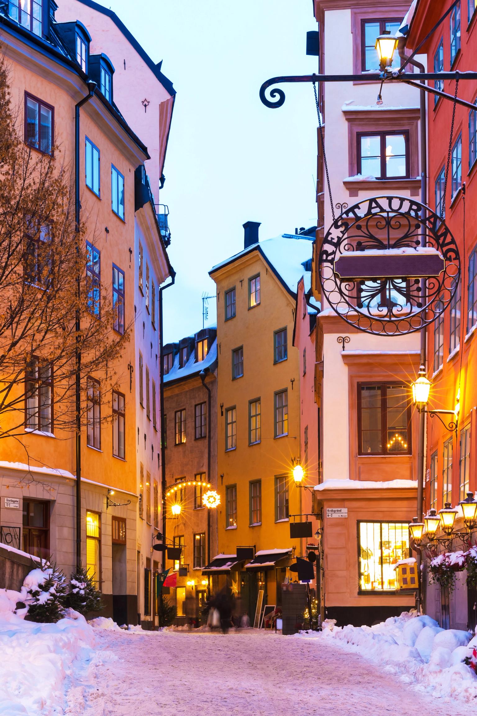 En snötäckt kullerstensgata i ett historiskt Stockholm, kantad med färgglada byggnader och lysande gatlyktor.