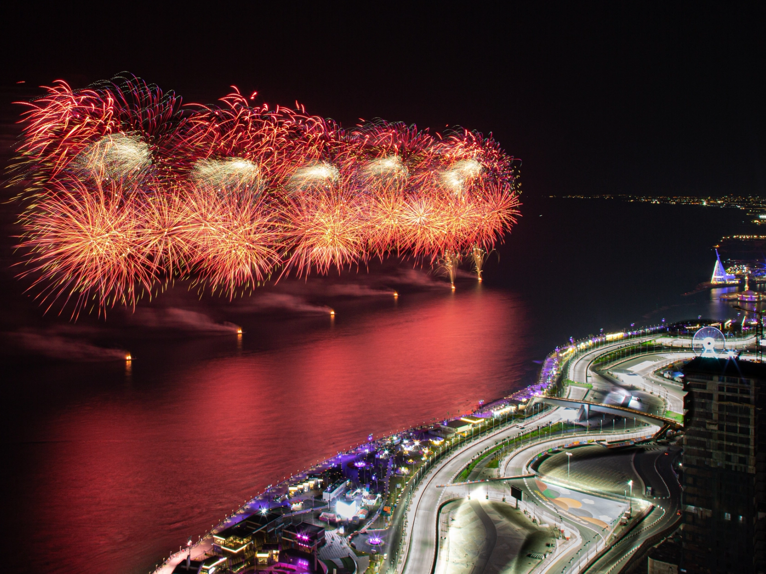 Rotes und goldenes Feuerwerk über einer hell erleuchteten stadt am Wasser in der Nacht.