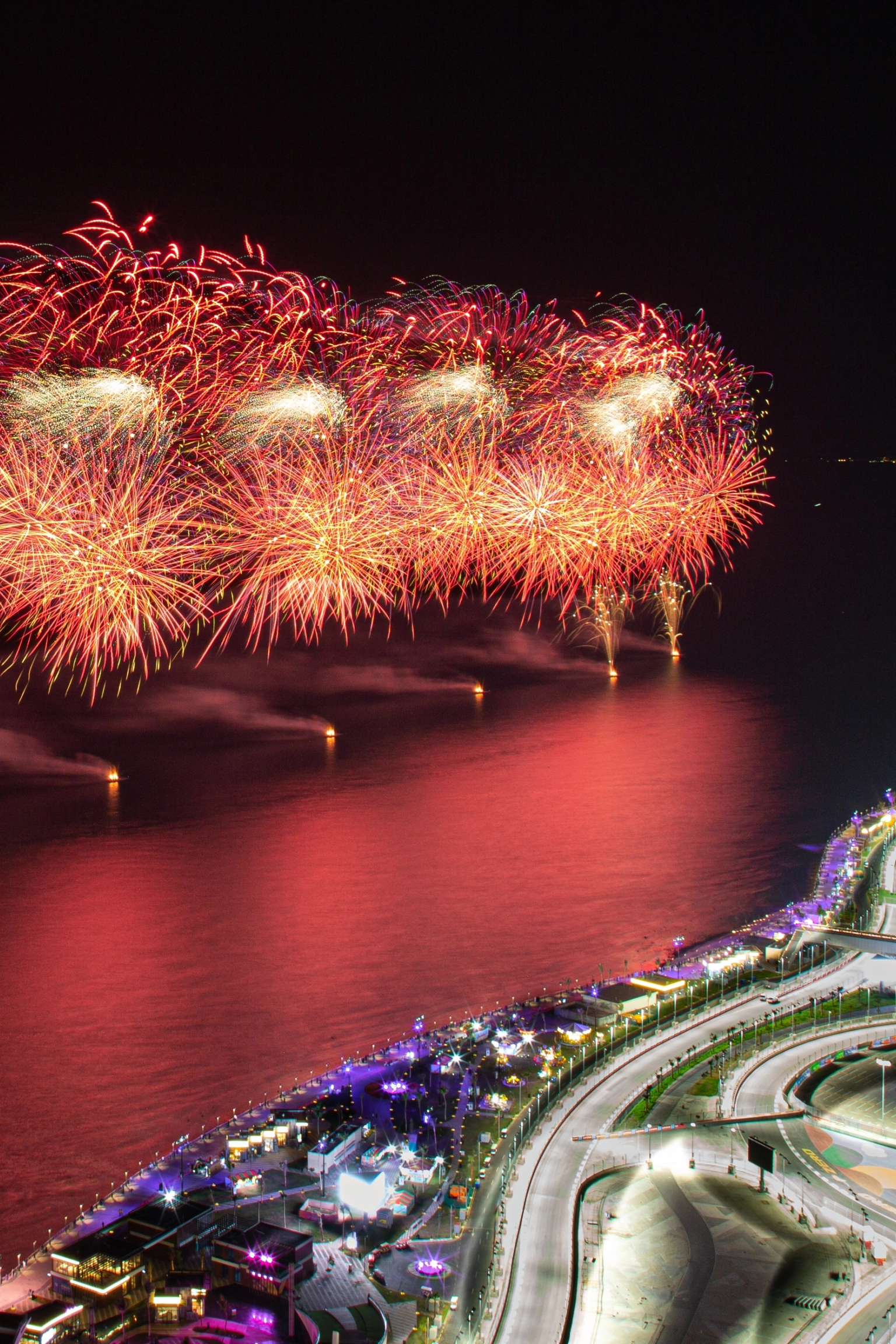 Rotes und goldenes Feuerwerk über einer hell erleuchteten stadt am Wasser in der Nacht.