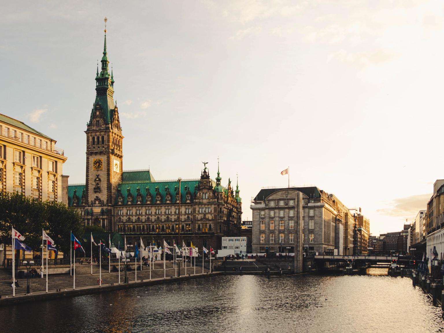 Ornate byggnad med ett högt klocktorn bredvid en kanal kantad med flaggor. Hamburg