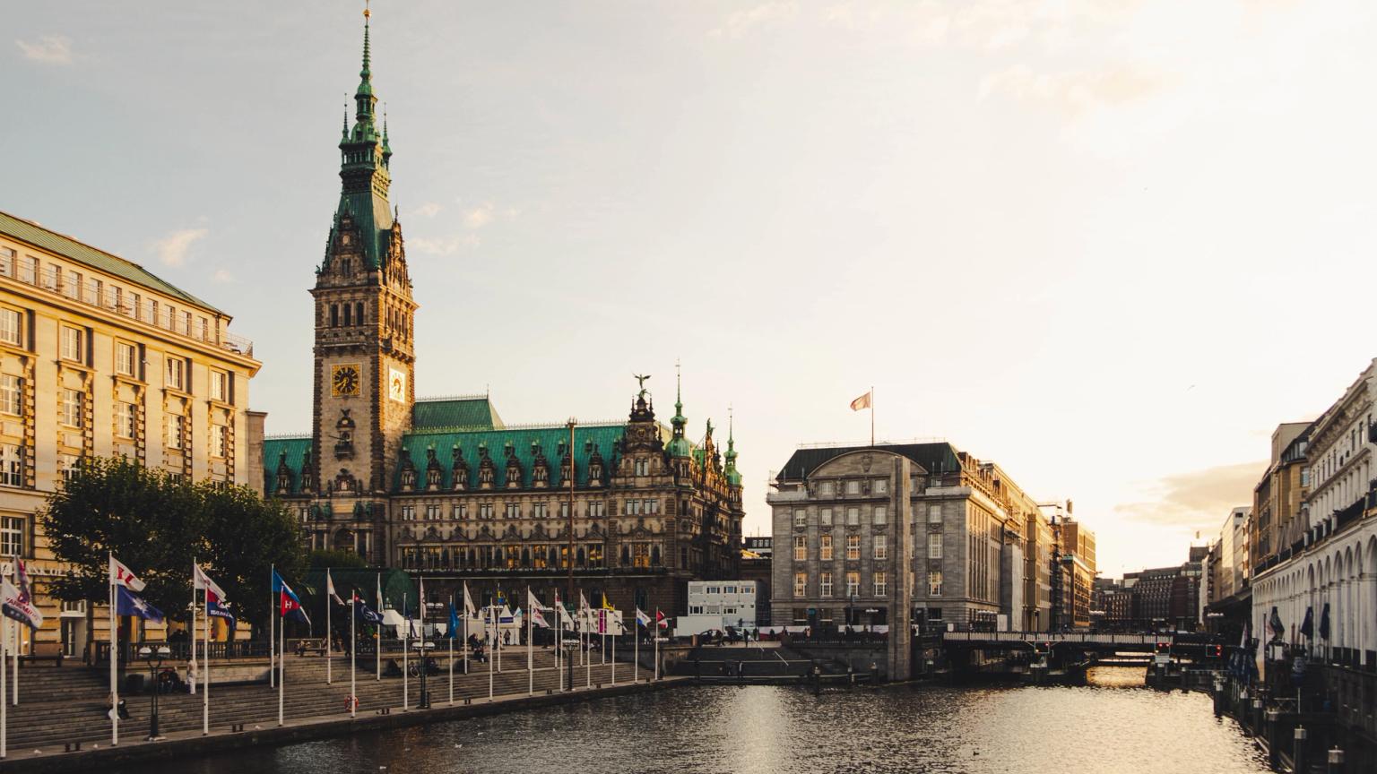Ornate byggnad med ett högt klocktorn bredvid en kanal kantad med flaggor. Hamburg
