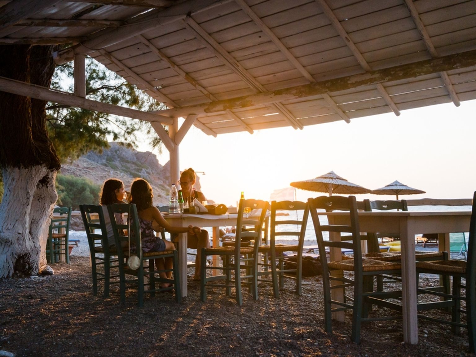 Tre personer äter på en rustik strandtaverna vid solnedgången.