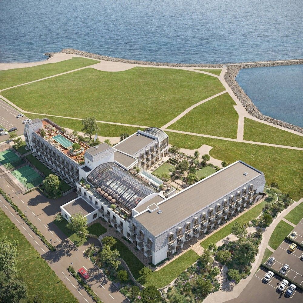 Flygvy över ett modernt hotellkomplex med en stor byggnad med glastakatrium, takpool, tennisbanor och anlagda grönområden intill havet. Rox Resort