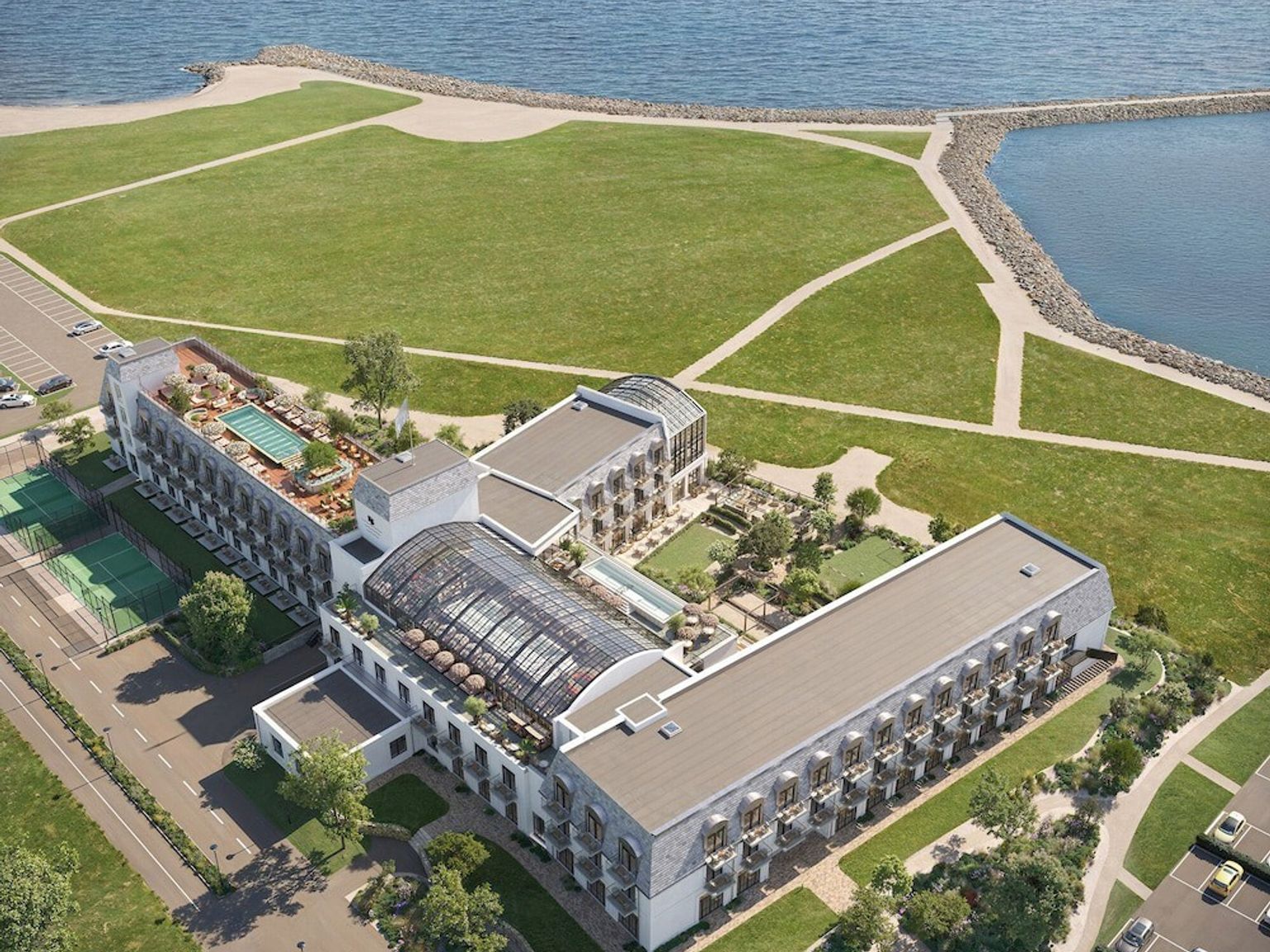 Flygvy över ett modernt hotellkomplex med en stor byggnad med glastakatrium, takpool, tennisbanor och anlagda grönområden intill havet. Rox Resort