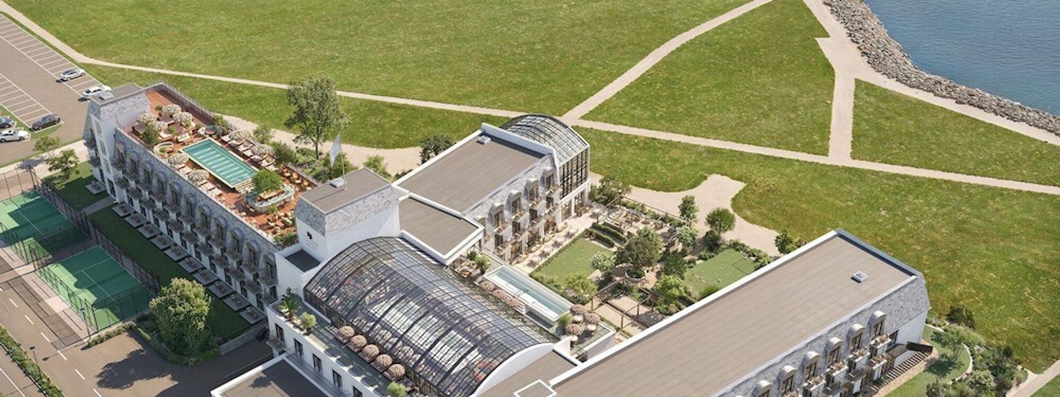 Flygvy över ett modernt hotellkomplex med en stor byggnad med glastakatrium, takpool, tennisbanor och anlagda grönområden intill havet. Rox Resort