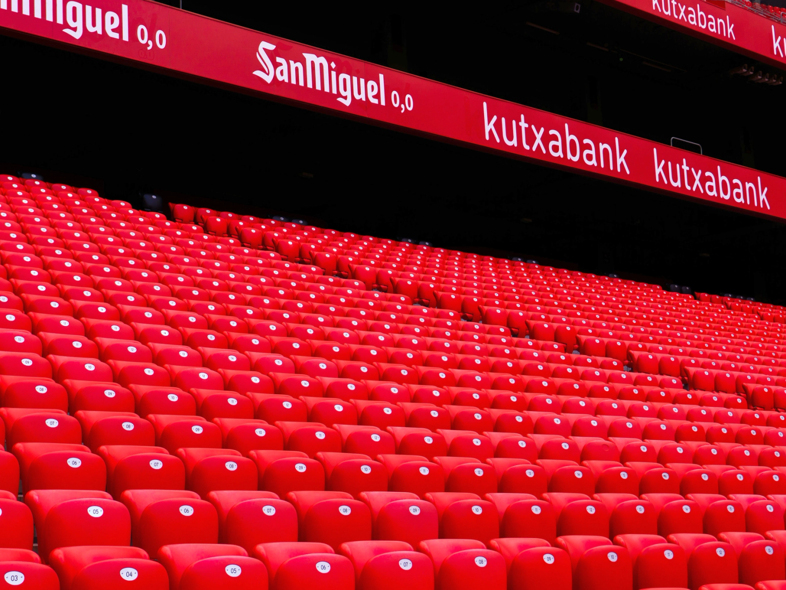 Rader av tomma röda stadionstolar med reklamskärmar ovanför.