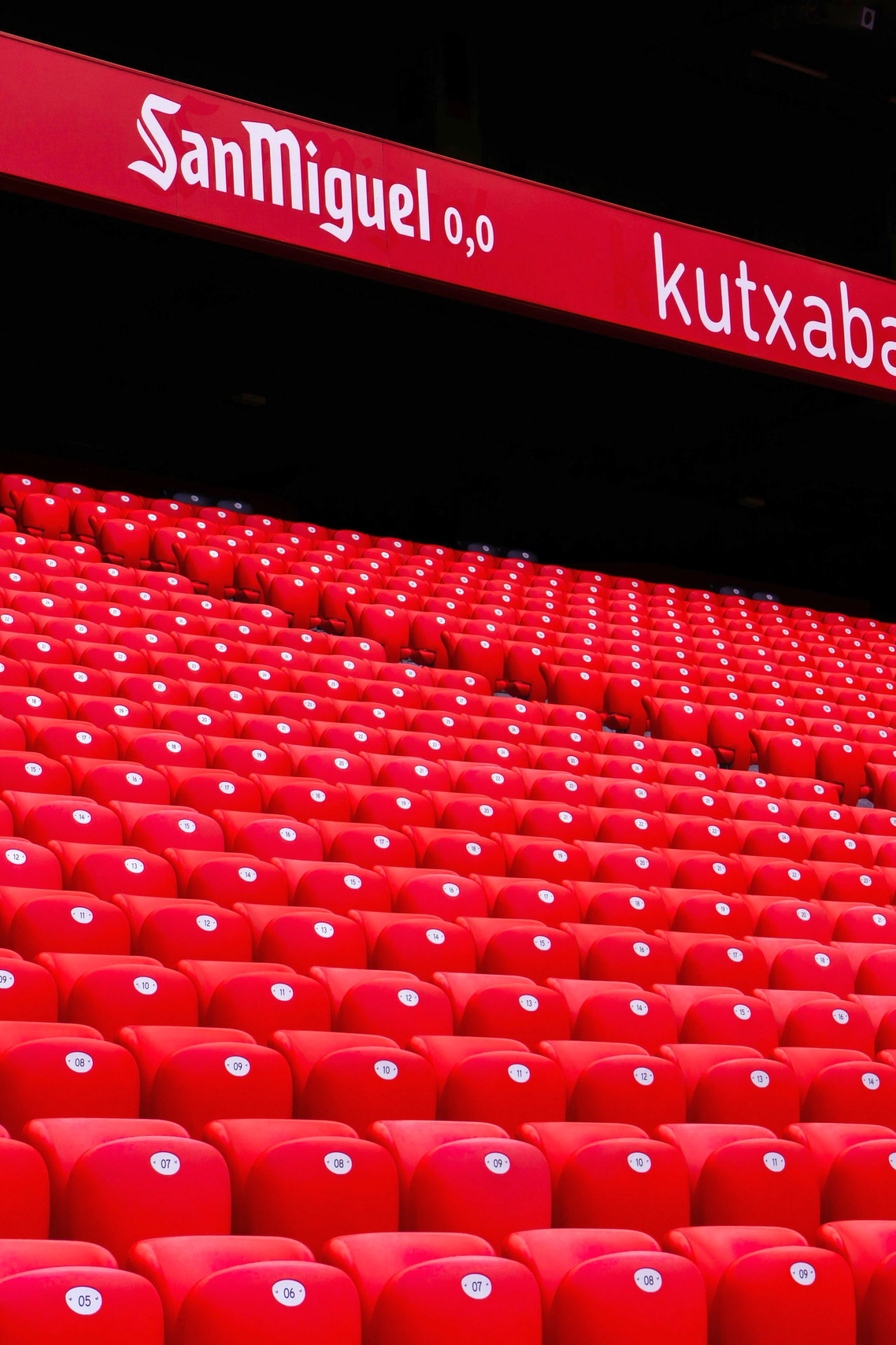 Rader av tomma röda stadionstolar med reklamskärmar ovanför.