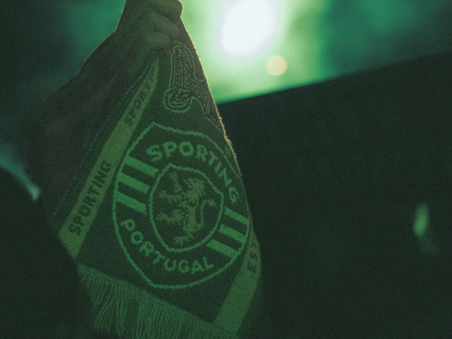 En hånd holder et grønt Sporting Portugal tørklæde med et løveemblem mod en mørk, grøn belyst baggrund.