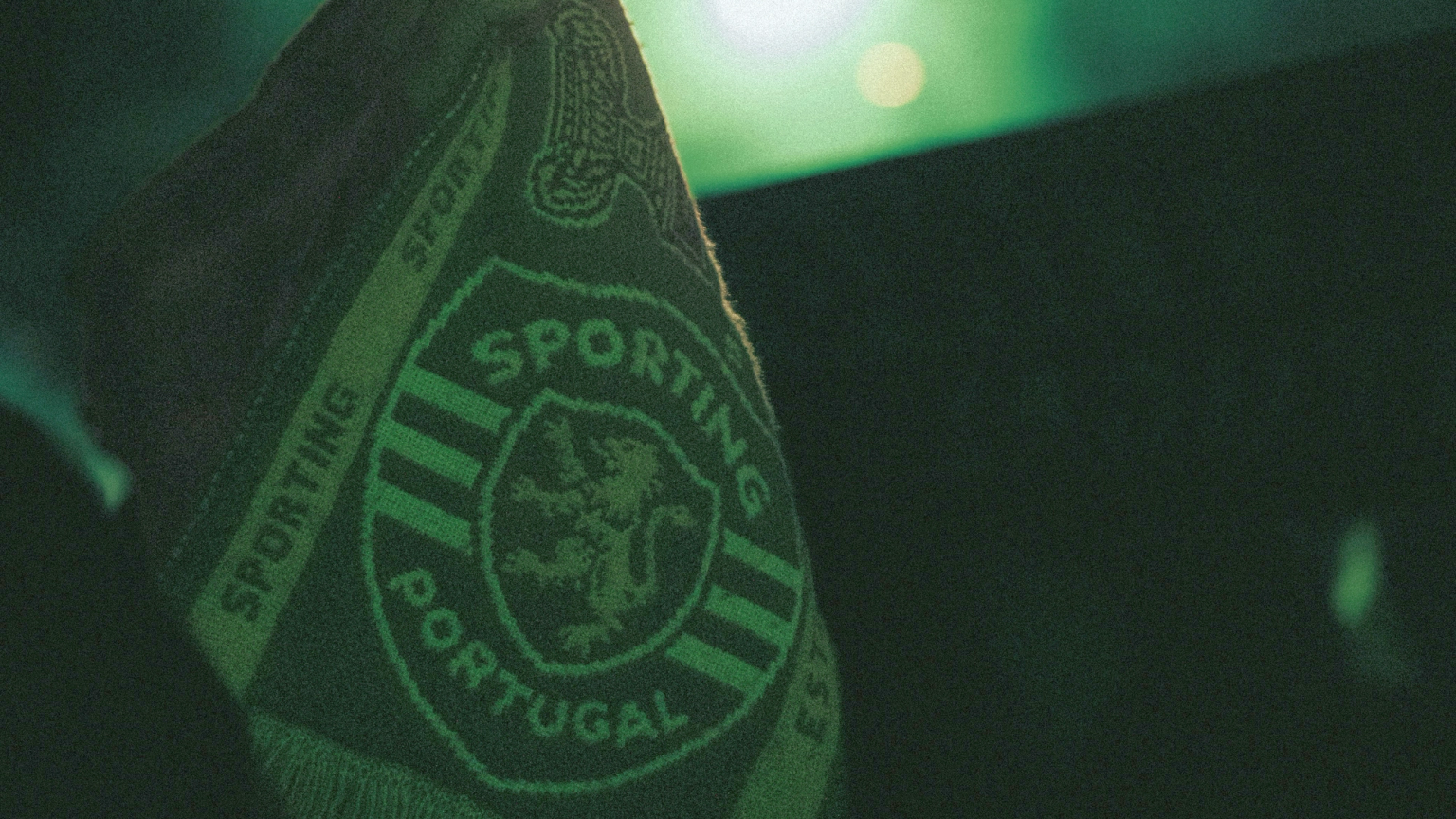 A hand holds up a green Sporting Portugal scarf with a lion crest against a dark, green-lit background.
