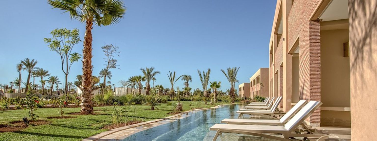 En solfylt feriestedscene med et langt svømmebasseng, solsenger, en bygning med balkonger, palmer og grønn plen under en klar blå himmel.
