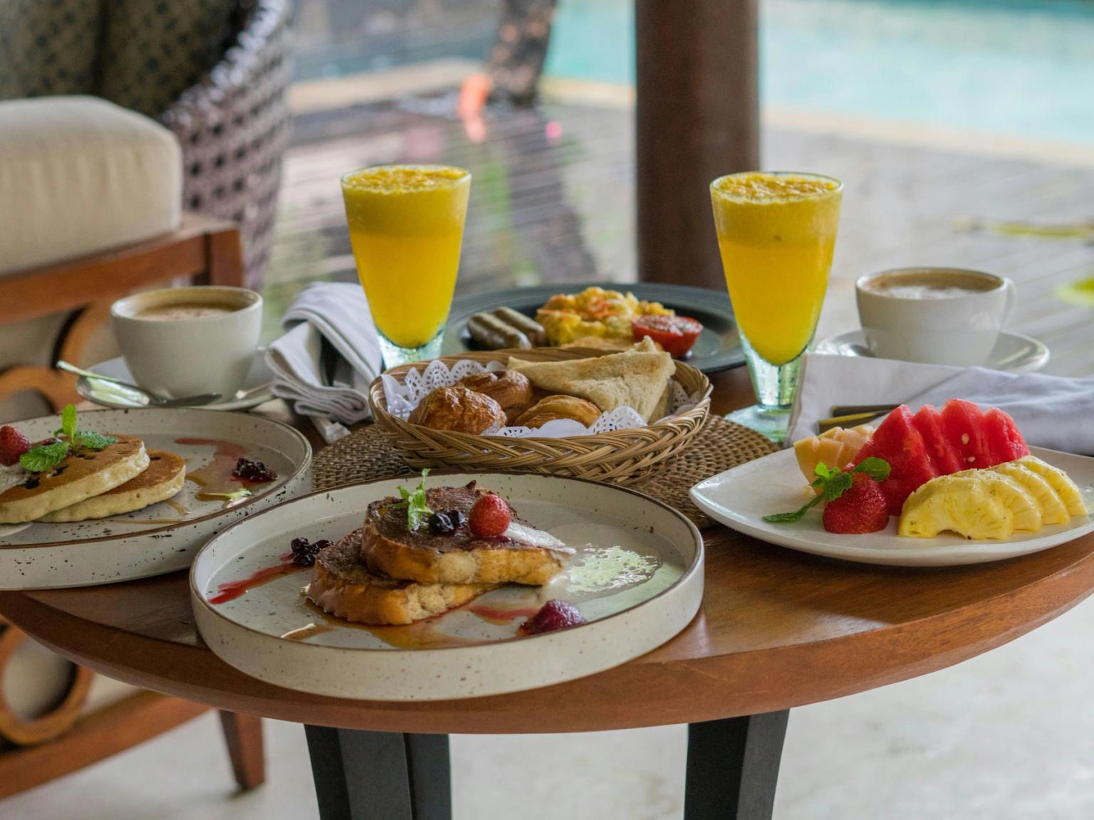 Et træbord udendørs ved en pool er dækket med en overdådig morgenmad med pandekager, french toast, frisk frugt, wienerbrød, kaffe og juice.