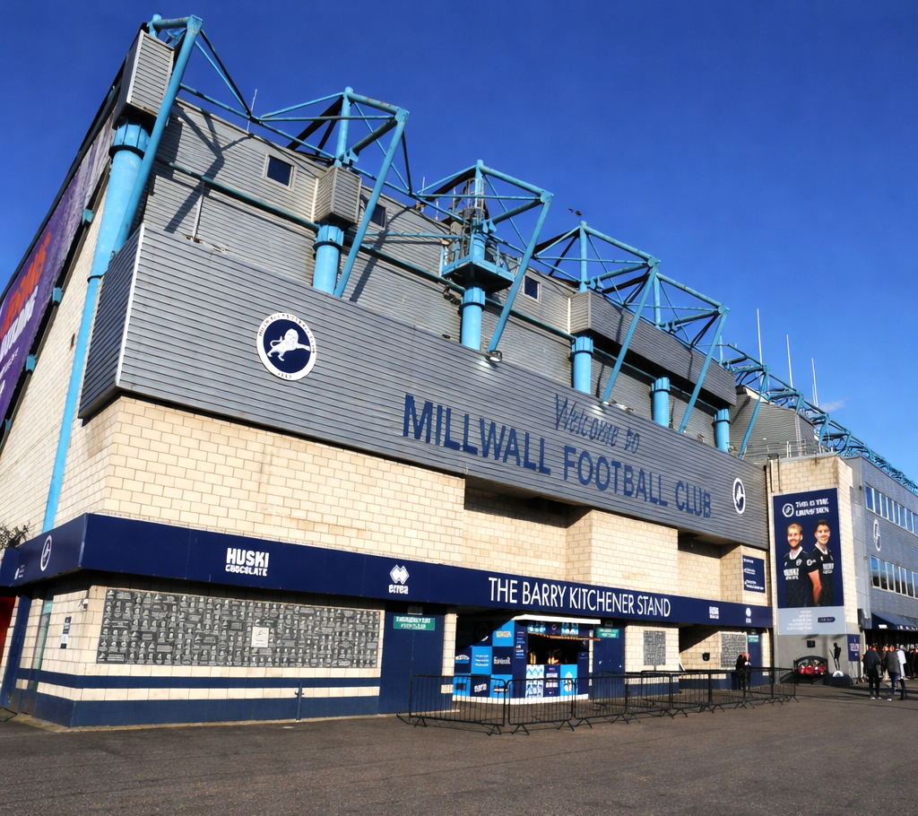 Vista exterior do estádio do Millwall Football Club, mostrando a entrada principal com os letreiros "Welcome to Millwall Football Club" e "The Barry Kitchener Stand".
