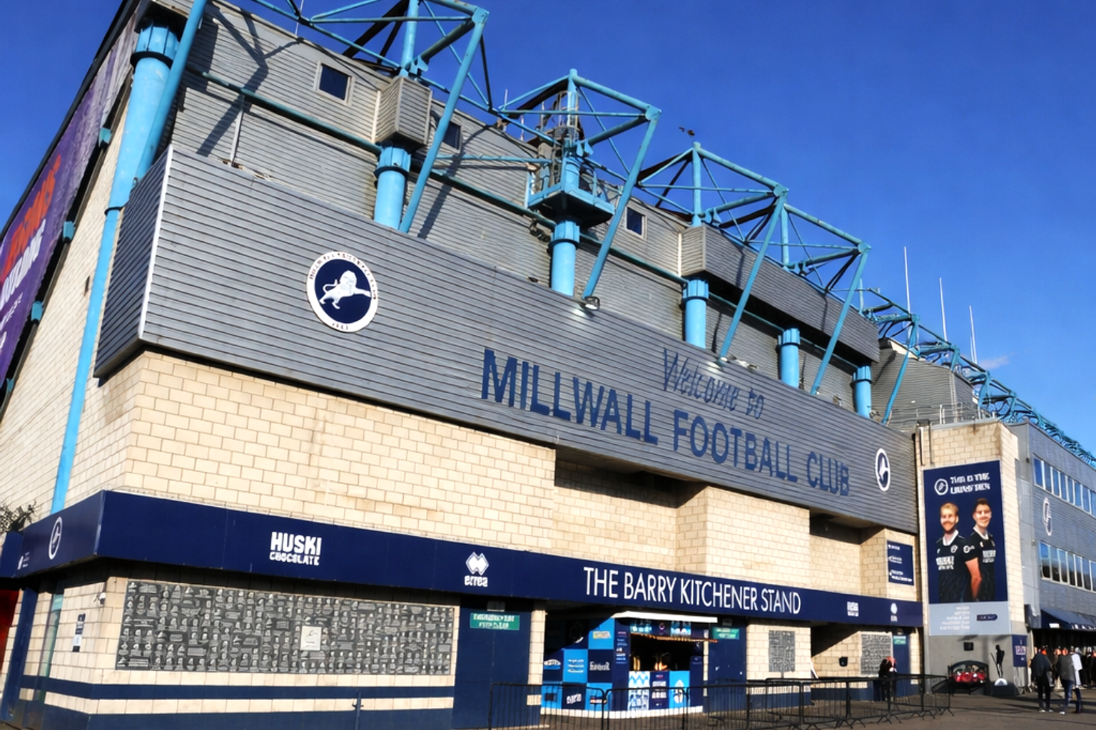 Buitenaanzicht van het Millwall Football Club stadion, met de hoofdingang en de borden "Welcome to Millwall Football Club" en "The Barry Kitchener Stand".