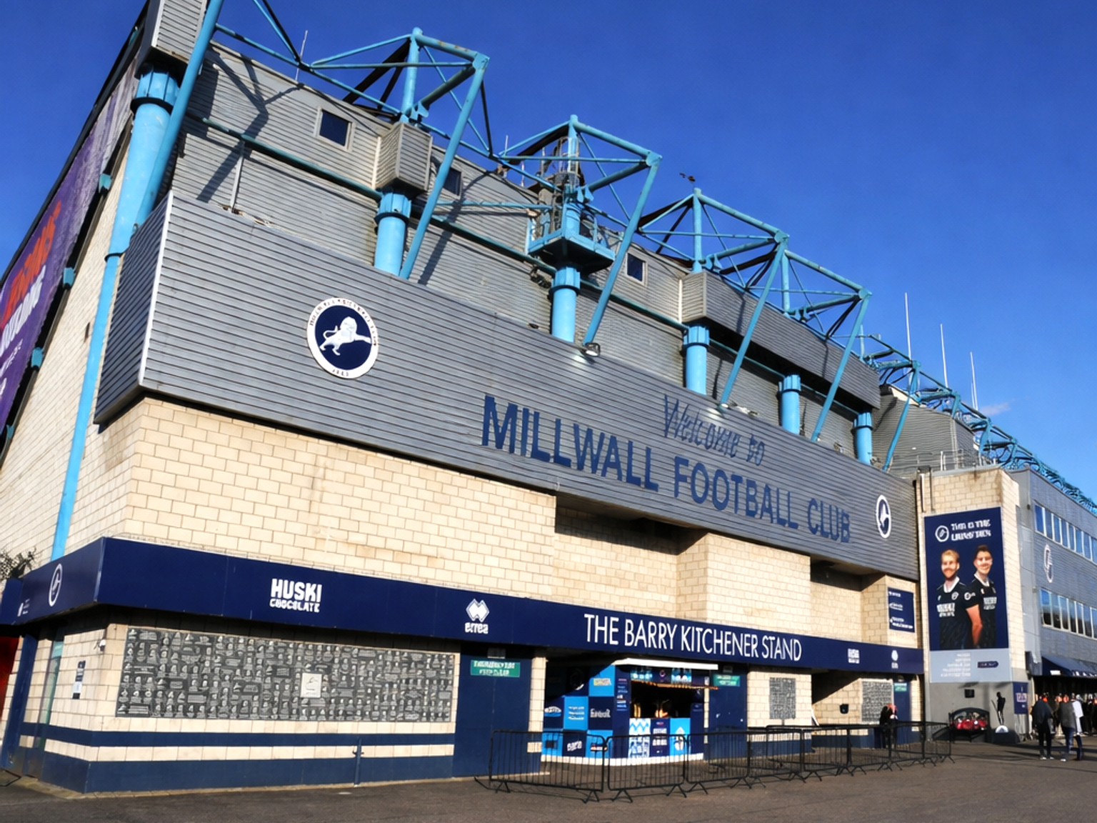 Vista exterior do estádio do Millwall Football Club, mostrando a entrada principal com os letreiros "Welcome to Millwall Football Club" e "The Barry Kitchener Stand".
