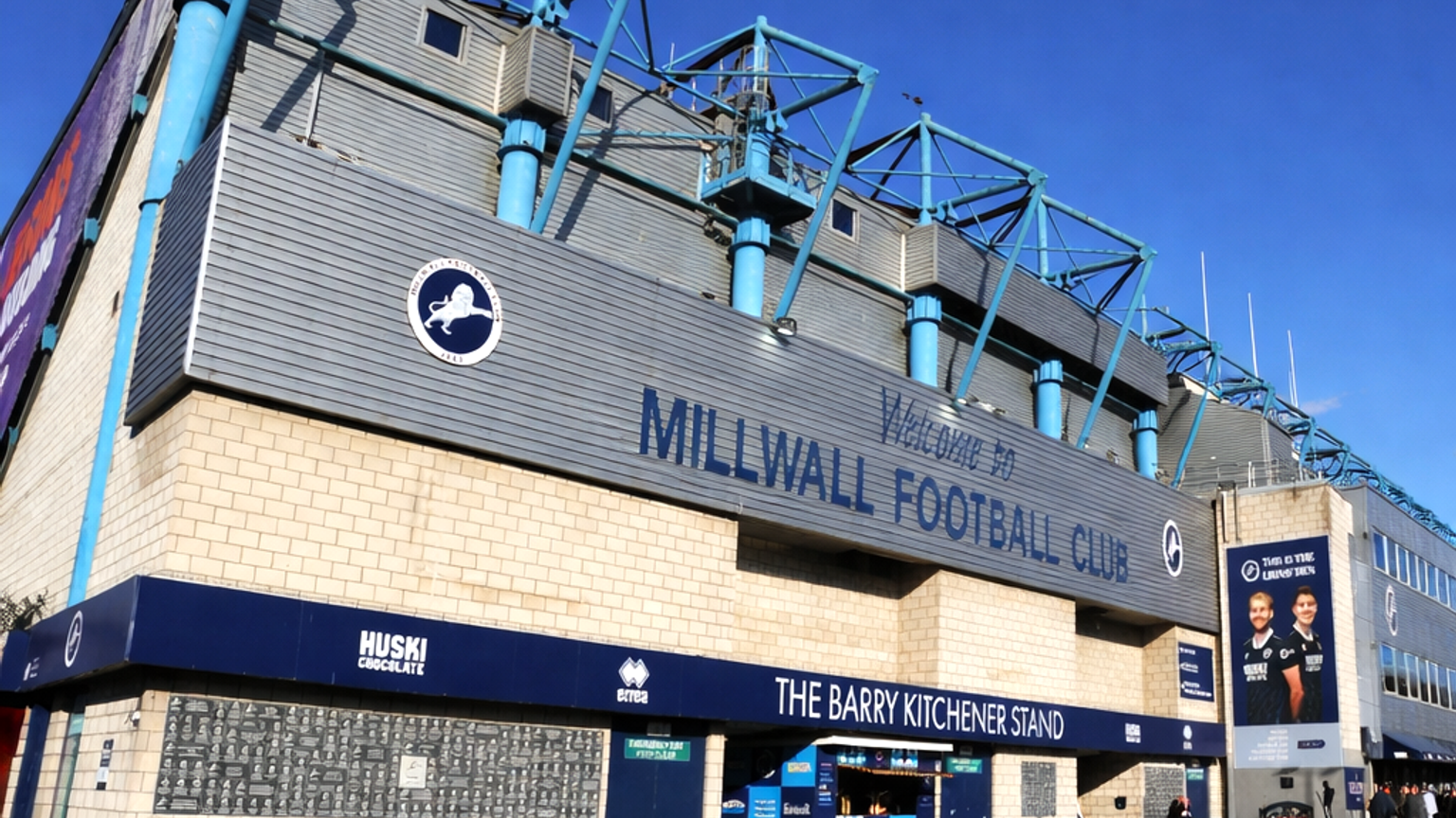 Buitenaanzicht van het Millwall Football Club stadion, met de hoofdingang en de borden "Welcome to Millwall Football Club" en "The Barry Kitchener Stand".