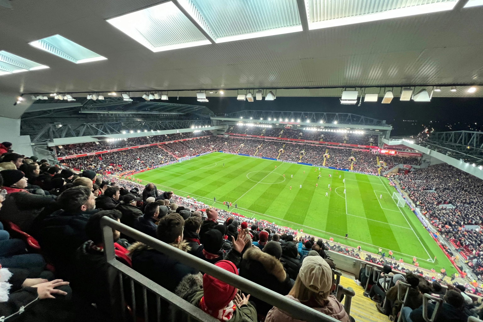 View from the upper stands of a packed football stadium at night, with a match being played on the illuminated green pitch below.