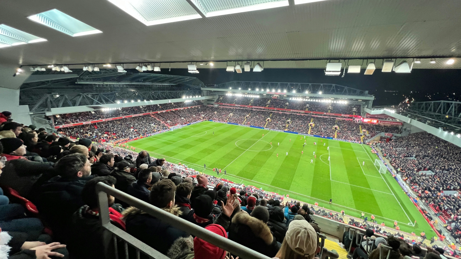 View from the upper stands of a packed football stadium at night, with a match being played on the illuminated green pitch below.
