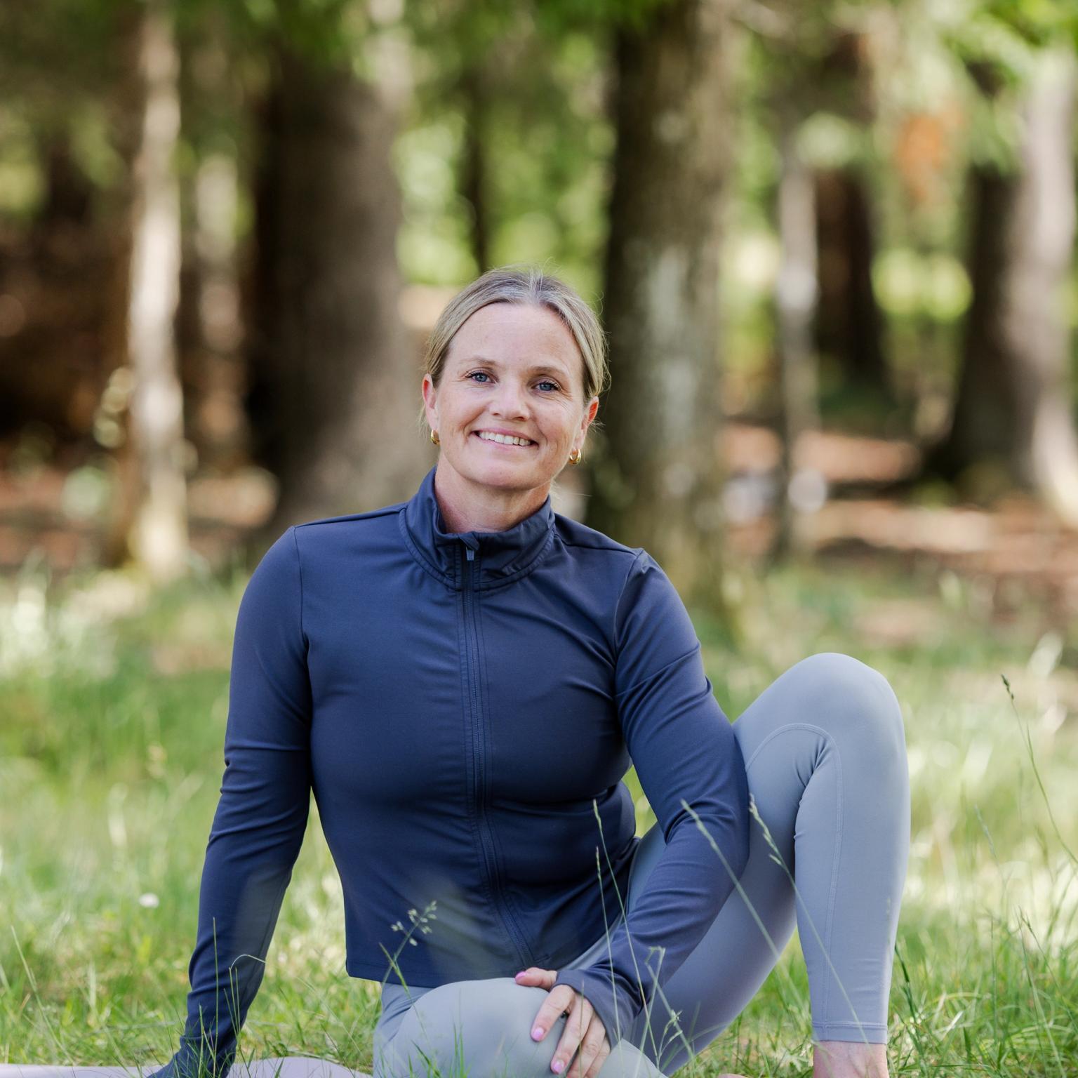 En leende kvinna i träningskläder som sitter i en yogaposition på en matta i en skog.