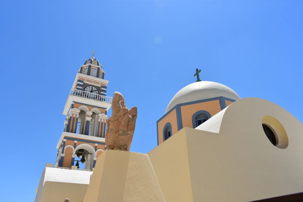 Klokketårn, kuppel og håndstatue af en kirke mod en klar blå himmel. Santorini, Grækenland