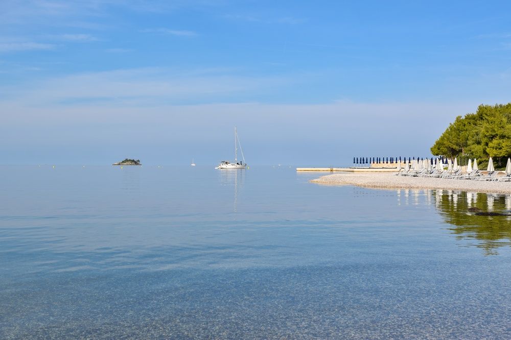 Klapperstensstrand med vita solstolar och blå parasoller vid ett klart blått hav, en avlägsen ö och en segelbåt. Lone Bay, Rovinj, Kroatien