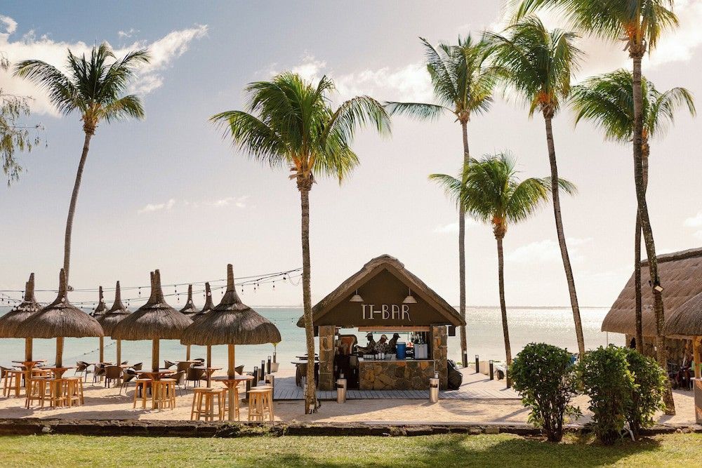 Ti-Baren, palmer och halmtakförsedda parasoller på en sandstrand vid havet.