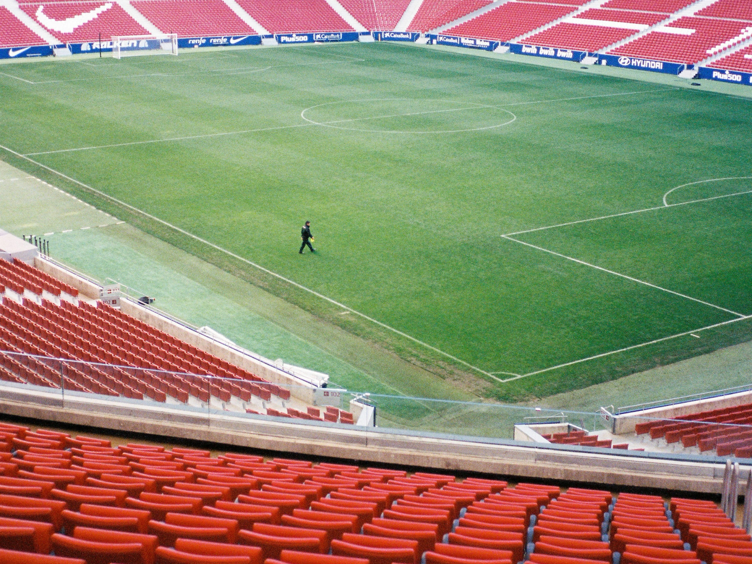 An empty soccer stadium, with rows of red seats surrounding a green field where a single person walks. "ATLETI" is spelled in white seats in the upper stands.