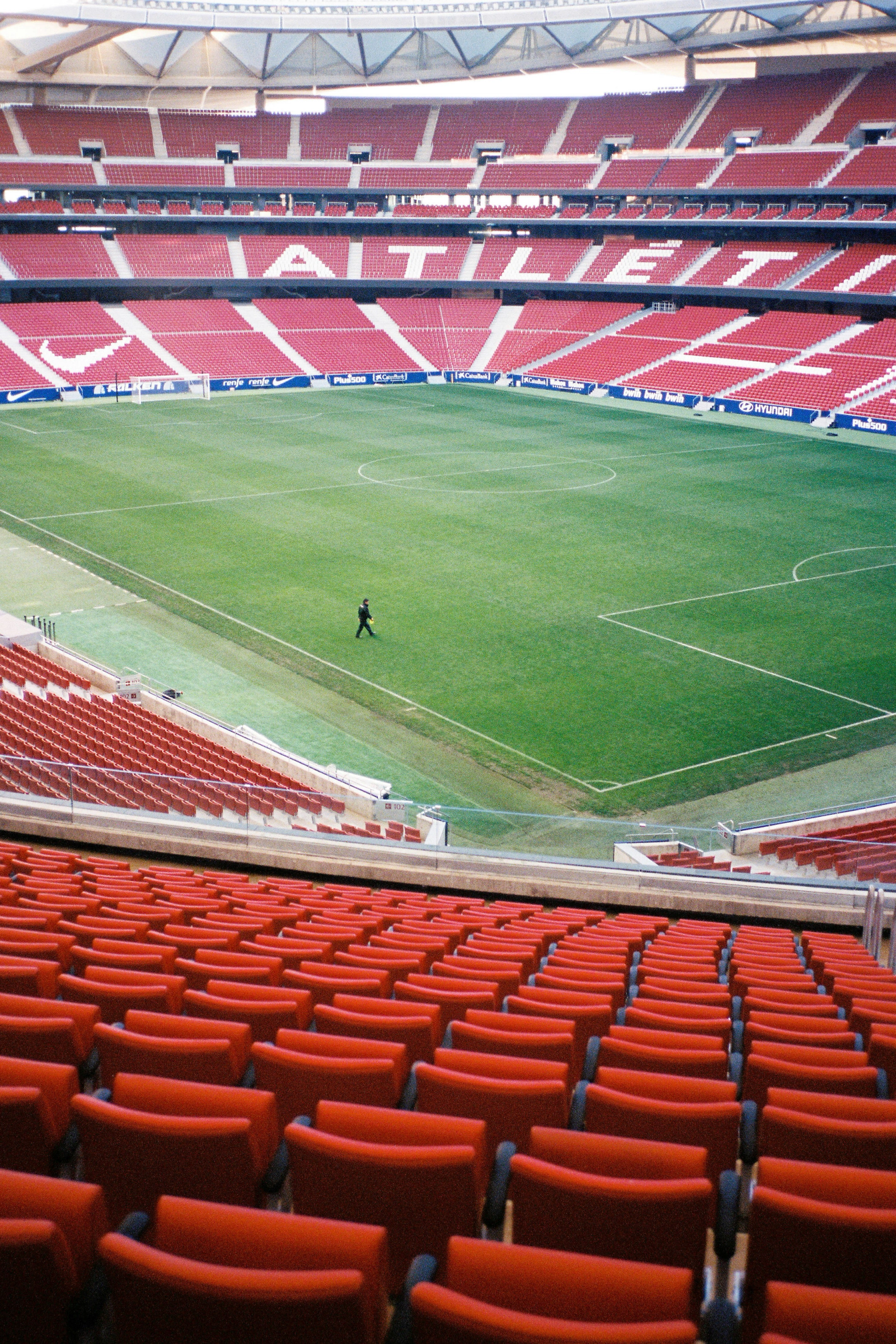An empty soccer stadium, with rows of red seats surrounding a green field where a single person walks. "ATLETI" is spelled in white seats in the upper stands.