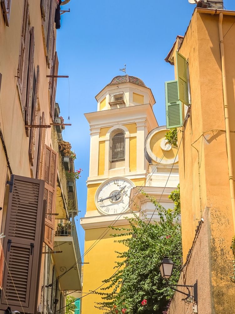 Ett gult klocktorn med vit urtavla och kupoltak reser sig mellan färgglada byggnader med öppna fönsterluckor och gröna växter, under en klarblå himmel. Villefranche-sur-Mer, Frankrike