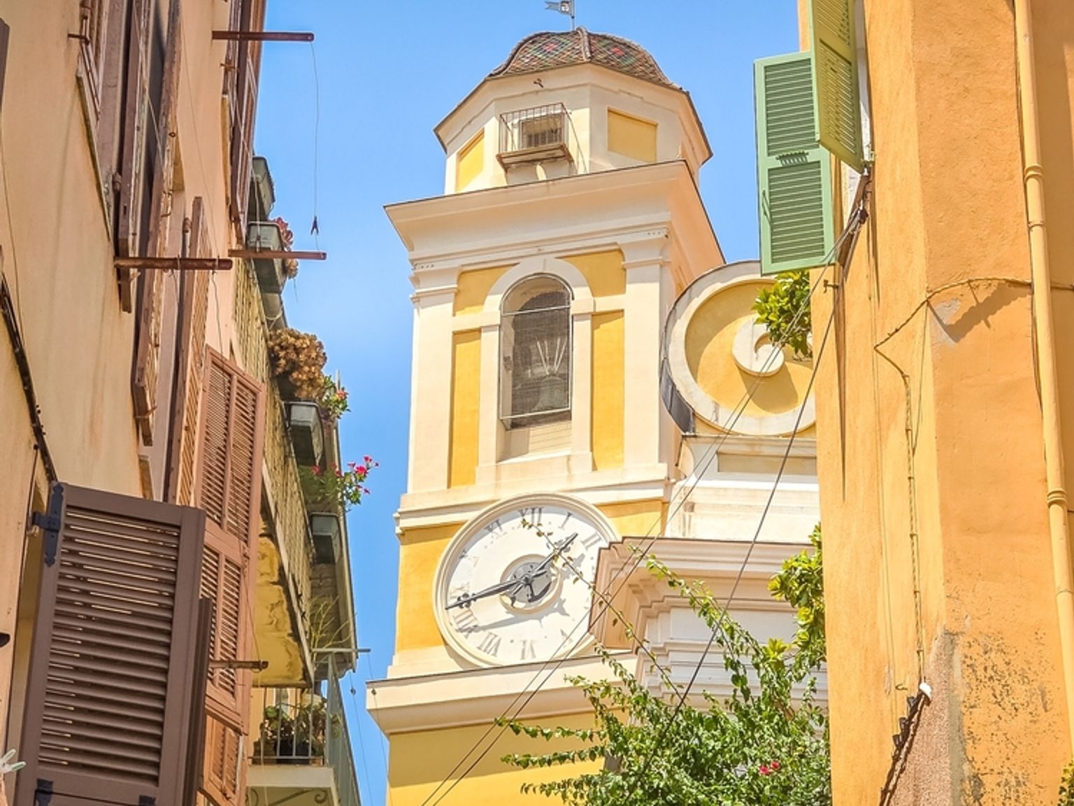 Keltainen kellotorni, jossa on valkoinen kellotaulu ja kupolikatto, kohoaa värikkäiden rakennusten välissä, joissa on avoimet ikkunaluukut ja vihreitä kasveja, kirkkaan sinisen taivaan alla. Villefranche-sur-Mer, Ranska