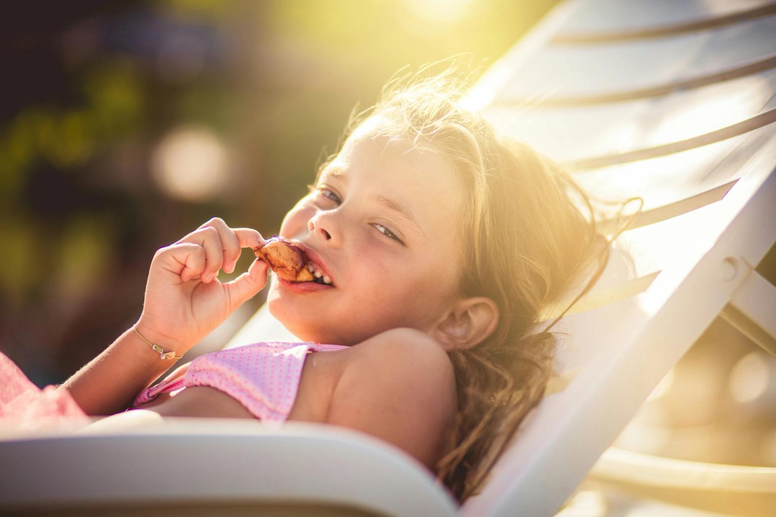 En ung flicka i en rosa baddräkt ligger på en solstol och äter glatt ett mellanmål i solen.