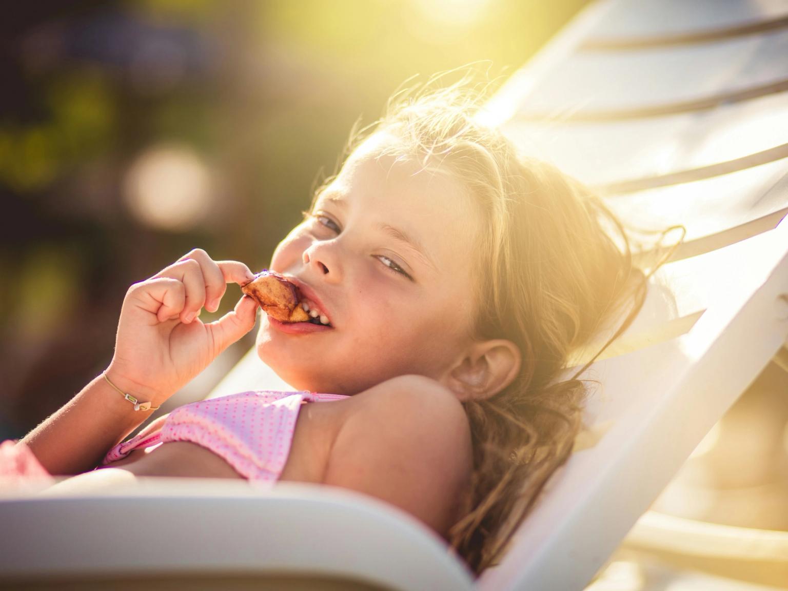 En ung flicka i en rosa baddräkt ligger på en solstol och äter glatt ett mellanmål i solen.