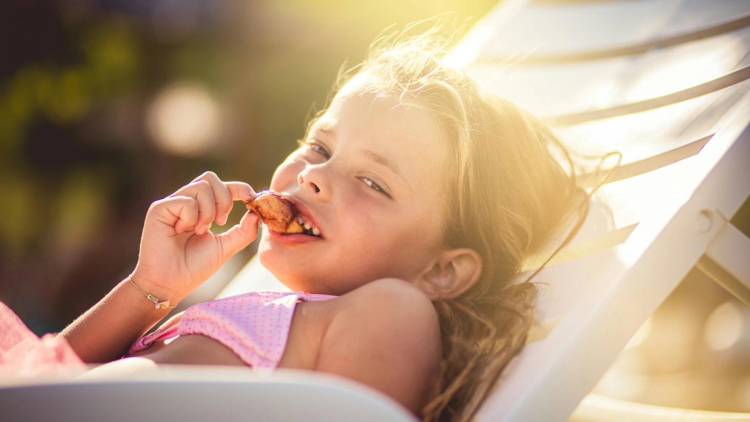 En ung flicka i en rosa baddräkt ligger på en solstol och äter glatt ett mellanmål i solen.
